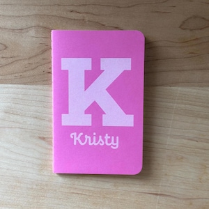 Op de afbeelding: Een felroze notitieboek met de witte letter "K" en de naam "Kristy" in sierlijke letters. Het rechthoekige notitieboek heeft afgeronde hoeken.