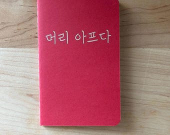 May include: A red notebook with the Korean text "머리 아프다" printed in white. The notebook is rectangular and appears to be closed. The background is a light brown wooden surface.
