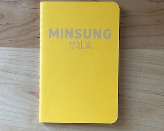 May include: A bright yellow notebook with rounded corners. The cover features the name "MINSUNG" in silver lettering, along with Korean characters. The notebook is lying on a light brown wooden surface.