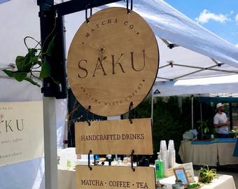May include: A hanging wooden sign display for Saku Matcha Co. featuring a circular top sign with the brand name and flower icon, followed by two rectangular signs reading Handcrafted Drinks and Matcha, Coffee, Tea.