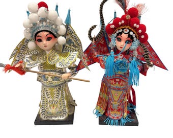 May include: Two Chinese opera dolls in elaborate costumes. One doll wears a white and gold outfit with a tall headdress and holds a spear. The other doll is in a red and gold costume with a large headdress and blue tassels.
