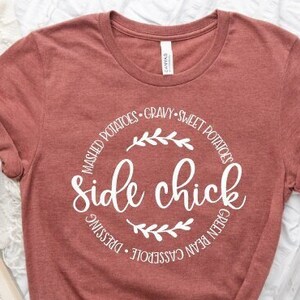 May include: A maroon t-shirt with a white circle graphic that says "side chick" surrounded by the words "mashed potatoes, gravy, sweet potatoes, green bean casserole, dressing".