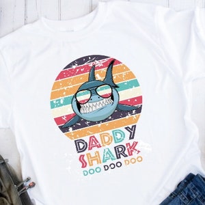 May include: White t-shirt with a graphic of a shark wearing sunglasses in front of a retro sunset. The text "Daddy Shark Doo Doo Doo" is printed below the shark.