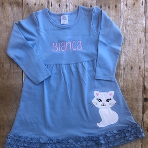 May include: A light blue long-sleeved dress with a white cat applique and a ruffled hem. The dress is personalized with the name "Bianca" in pink.