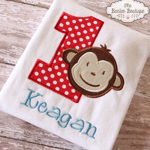 May include: A white t-shirt with a red and white polka dot number one and a brown monkey face. The name "Keagan" is embroidered in blue below the number.
