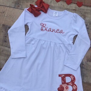 May include: White long-sleeved dress with a ruffled hem. The dress has a red embroidered initial "B" with hearts and a red embroidered name "Bianca".
