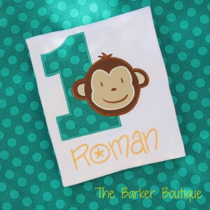 May include: A white onesie with a green and white polka dot number one, a brown monkey face, and the name "Roman" in yellow with a yellow star. The text "The Barker Boutique" is at the bottom of the onesie.