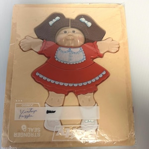 May include: Vintage Cabbage Patch Kids jumbo puzzle in a plastic bag. The puzzle is of a girl with brown hair in pigtails wearing a red dress with white trim and white shoes.