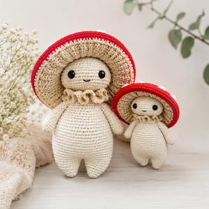 May include: Two handmade crochet mushroom dolls with cream-colored bodies, ruffled collars, and wide red caps featuring white spots. The dolls have black bead eyes and simple stitched smiles. The larger doll stands next to a smaller version.