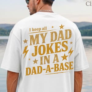 Könnte beinhalten: Weißes T-Shirt mit goldenem Text: "I keep all MY DAD JOKES IN A DAD-A-BASE". Die Person trägt eine blaue Kappe und Sonnenbrille. Im Hintergrund ein See und Berge, sowie das Logo ClimbingRose Artisan.