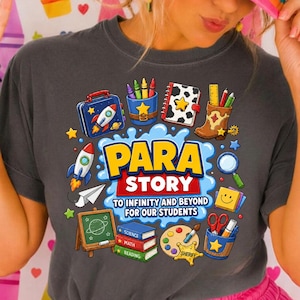 Puede incluir: Una camiseta gris oscuro con un gráfico colorido de útiles escolares que incluye un cohete, libros, una pizarra y una paleta de pintura. El texto dice PARA STORY TO INFINITY AND BEYOND FOR OUR STUDENTS. Se lleva una gorra con la palabra TEACHER.