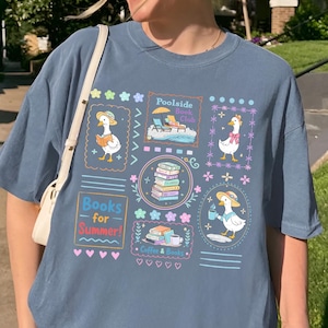 May include: A blue jean colored t-shirt with a graphic design featuring cartoon ducks, books, and the text "Books for Summer!". The design includes the word "Poolside" and other summer-themed illustrations.