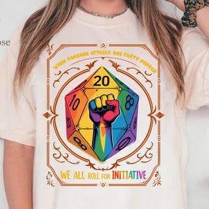 May include: Ivory-colored t-shirt featuring a colorful die design with a clenched fist and the text "WHEN SOMEONE ATTACKS ONE PARTY MEMBER WE ALL ROLL FOR INITIATIVE". The design includes the numbers 8, 10, and 20.