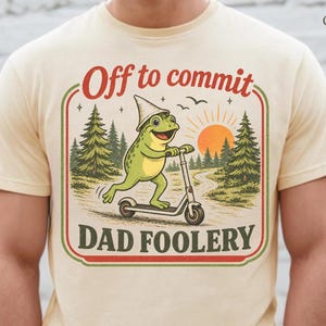 Puede incluir: Camiseta color crema con un diseño gráfico de una rana en patinete, con el texto "Off to commit Dad Foolery" en un marco rojo y verde. El diseño incluye un atardecer y árboles.