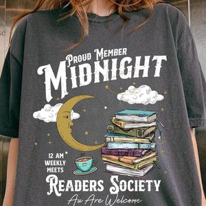 Könnte beinhalten: Dunkelgraues T-Shirt mit einem Design für einen Buchclub. Das Design enthält den Text "Proud Member Midnight Readers Society" mit einem Mond, Wolken, Büchern und einer Teetasse. Das Shirt ist ein lässiger, bequemer Stil.