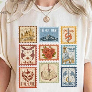 Op de afbeelding: Crèmekleurig T-shirt met negen postzegel-achtige afbeeldingen. De postzegels tonen verschillende illustraties, waaronder een berg, rozen en een zwaard. Tekst op het shirt is onder meer "LUNATHION", "THE NIGHT COURT", "STAR BORN", "the Bat Boys", "FIREHEART" en "UMBRA MORTIS".