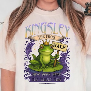 May include: Cream-coloured t-shirt with a cartoon frog wearing a crown and holding a sign that says "HALP." The shirt has the text "KINGSLEY THE FROG" above the frog and "HE'S BEEN HERE THE WHOLE TIME" below.
