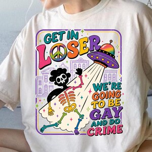 May include: Cream-colored t-shirt with a colorful graphic. The graphic features a skeleton being beamed up by a UFO, with text that reads "GET IN LOSER" and "WE'RE GOING TO BE GAY AND DO CRIME."