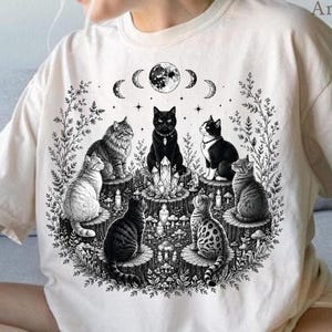 May include: A cream-colored t-shirt featuring a black ink graphic of six cats sitting on tree stumps in a circle around a central crystal. The design includes a full moon, crescent moon phases, stars, and foliage. The text ClimbingRose Artisan is visible.