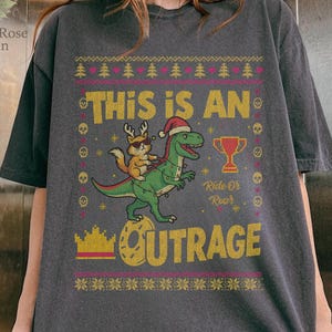 May include: Dark gray t-shirt with a festive design. The shirt features a green dinosaur wearing a Santa hat, ridden by a fox with antlers. Text includes "THIS IS AN OUTRAGE" and "Ride Or Roar".