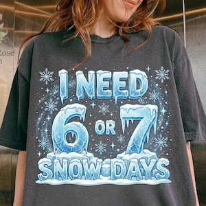May include: Dark gray t-shirt with the text "I NEED 6 OR 7 SNOW DAYS" in icy blue lettering. The design includes snowflakes and ice accents, creating a winter theme. The shirt is a casual wear item.