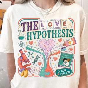 以下が含まれることがあります： 「THE LOVE HYPOTHESIS」というテキストがプリントされたクリーム色のTシャツ。顕微鏡、試験管、ビーカー、DNA鎖、そして「The Love Hypothesis」というタイトルの本など、科学的な要素が描かれたカラフルなグラフィックデザインが特徴です。