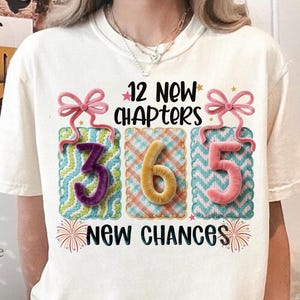Könnte beinhalten: Cremefarbenes T-Shirt mit dem Text "12 New Chapters 365 New Chances". Das Design zeigt drei Geschenkbox-Grafiken mit den Zahlen 3, 6 und 5 in Lila, Gelb und Rosa. Rosa Schleifen und Feuerwerk vervollständigen das Design.