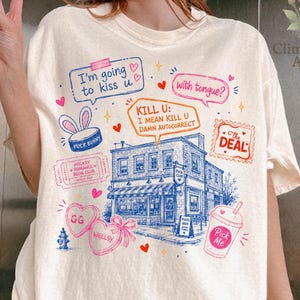 May include: Off-white t-shirt featuring a playful graphic with speech bubbles and quirky phrases. The design includes a building illustration and various stamps and hearts. The shirt has a loose fit and is made of a soft material.