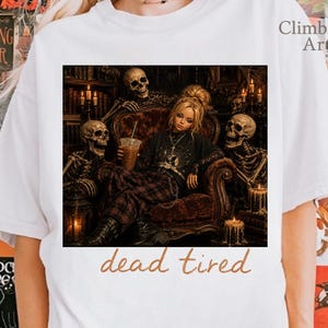 May include: A white short-sleeve t-shirt featuring a graphic print of a person sitting in a dark velvet chair surrounded by three skeletons in a dimly lit room. The person holds a beverage. The text "dead tired" is printed below the graphic.