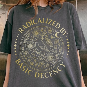 Könnte beinhalten: Dunkelgraues T-Shirt mit goldenem, kreisförmigem Design, das einen Vogel, Blumen und den Text "RADICALIZED BY BASIC DECENCY" zeigt. Das Shirt hat eine lockere Passform und kurze Ärmel.