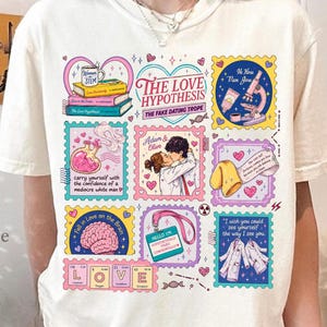 以下が含まれることがあります： The Love Hypothesisにインスパイアされたカラフルなイラストのグリッドが描かれた、クリーム色の半袖Tシャツ。グラフィックには、本、顕微鏡、脳、カップル、白衣、そしてThe Love Hypothesisなどのテキスト要素が含まれています。