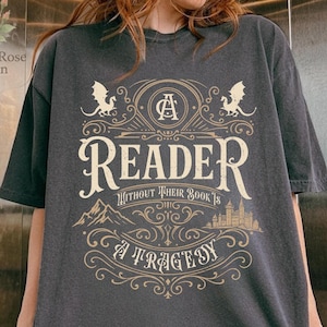 Może przedstawiać: Ciemnoszara koszulka z napisem "A Reader Without Their Book's A Tragedy" w stylu vintage. Projekt zawiera smoki, góry i zamek. Koszulka ma luźny, obszerny krój.