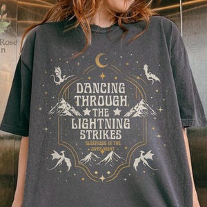 Pode incluir: Camiseta cinza escuro com um design gráfico com o texto "Dancing Through The Lightning Strikes" e ilustrações de montanhas, dragões, uma lua e estrelas. A camiseta tem um corte folgado e mangas curtas.