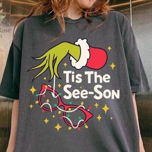 May include: Dark gray t-shirt featuring a graphic of a green hand, red glove, and red glasses with Christmas lights. The text "Tis The See-Son" is printed on the shirt. The shirt is a casual, oversized style.