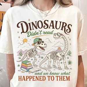May include: Cream-colored t-shirt featuring a humorous graphic of dinosaur skeletons wearing sunglasses and a hat, with the text "Dinosaurs Didn't Read and we know what HAPPENED TO THEM". The design includes books, a sun, and desert scenery.