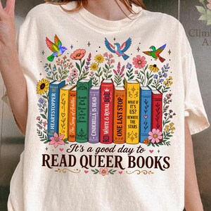Op de afbeelding: Crèmekleurig T-shirt met een kleurrijke afbeelding van boeken, bloemen en vogels. De tekst "It's a good day to READ QUEER BOOKS" staat onder de boeken. Het ontwerp bevat de namen van verschillende boeken.