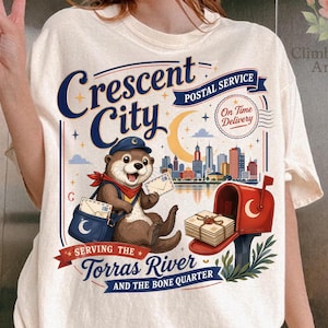 May include: Cream-colored t-shirt featuring a graphic of an otter in a postal worker uniform, holding letters. The design includes the text "Crescent City Postal Service" and "Serving the Torras River and the Bone Quarter."