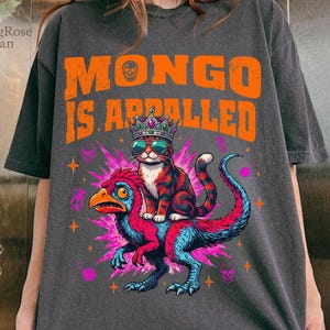 Può includere: T-shirt grigio scuro con la scritta "MONGO IS APPALLED" in arancione. La grafica presenta un gatto con occhiali da sole e una corona in sella a una creatura colorata simile a un dinosauro. Il design include stelle e teschi.