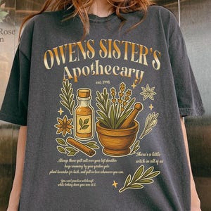以下が含まれることがあります： 「OWEN'S SISTER'S Apothecary est. 1995」というテキストがプリントされたチャコールグレーのTシャツ。デザインには、乳鉢と乳棒、ガラス瓶、ハーブ、シナモンスティックが描かれており、小さな文字と星のアクセントが添えられています。