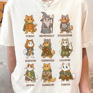 Puede incluir: Camiseta color crema con nueve gatos de dibujos animados disfrazados de personajes de fantasía. Cada gato tiene un disfraz único y un nombre, como "Furdo" y "Meowndalf". La camiseta tiene el texto "ClimbingRose Artisan" en el lado izquierdo.