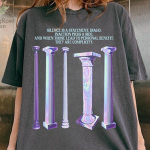 May include: Dark gray t-shirt featuring a graphic of five stylized columns in shades of purple and blue. The shirt has text that reads "SILENCE IS A STATEMENT. INACTION PICKS A SIDE. AND WHEN THOSE LEAD TO PERSONAL BENEFIT. THEY ARE COMPLICITY."