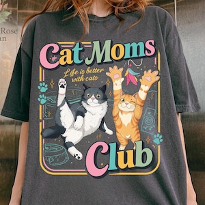 Op de afbeelding: Donkergrijs t-shirt met een kleurrijke afbeelding met de tekst "Cat Moms Club". Het ontwerp toont twee cartoonkatten, een zwart-witte, de andere oranje, met de tekst "Life is better with cats."