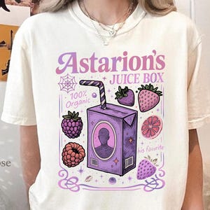 Puede incluir: Camiseta color crema con un diseño de caja de jugo púrpura con el texto "Astarion's Juice Box". El diseño incluye ilustraciones de bayas y una pajita. La camiseta también tiene el texto "100% Organic" y "his favorite".