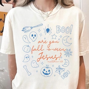 Puede incluir: Camiseta color crema con un diseño de Halloween. El diseño incluye ilustraciones azules y naranjas de fantasmas, calabazas, una araña y la frase "Boo! are you fall-o-ween Jesus?" con una referencia bíblica.