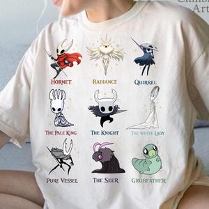 Puede incluir: Camiseta color crema con nueve ilustraciones de personajes del videojuego Hollow Knight. Cada personaje tiene un nombre impreso debajo, incluyendo Hornet, Radiance y Quirrel. El logotipo de ClimbingRose Artisan está en la esquina superior derecha.
