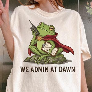 May include: A cream-colored t-shirt featuring a graphic of a green frog wearing a red cape, holding a walkie-talkie and a clipboard while sitting on a rock. The text below the frog reads WE ADMIN AT DAWN in bold, dark brown capital letters.