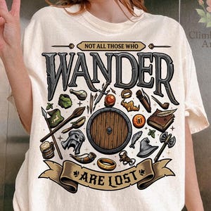 May include: Cream-colored t-shirt with a graphic design featuring the words "WANDER" and "NOT ALL THOSE WHO ARE LOST." The design includes a shield, sword, and other fantasy-themed illustrations. The shirt is a relaxed fit.