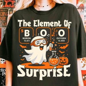 Pode incluir: Camiseta preta com um gráfico temático de Halloween. O design apresenta um fantasma usando óculos, segurando um tubo de ensaio e o texto "The Element Of BOO Surprise". Inclui uma abóbora e morcegos.