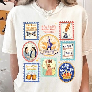 以下が含まれることがあります： クリーム色のTシャツ。カラフルな切手デザインのコラージュが特徴です。切手にはピストル、羽根ペンとインク壺の画像、そして「HAMILTON」の文字が含まれています。他の切手には「I AM NOT THROWING AWAY MY SHOT」と「You Want A Revolution」というフレーズがあります。