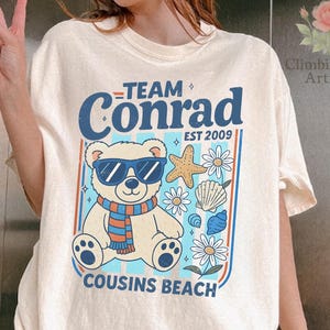 May include: Cream-colored t-shirt featuring a graphic of a bear wearing sunglasses and a scarf, surrounded by seashells and flowers. The text reads "Team Conrad EST 2009 Cousins Beach."